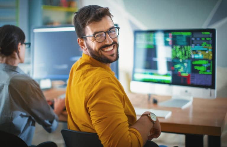 Entwickler sitzt lächelnd vor seinem geöffneten Arbeitsplatz mit geöffnetem Systemprogramm